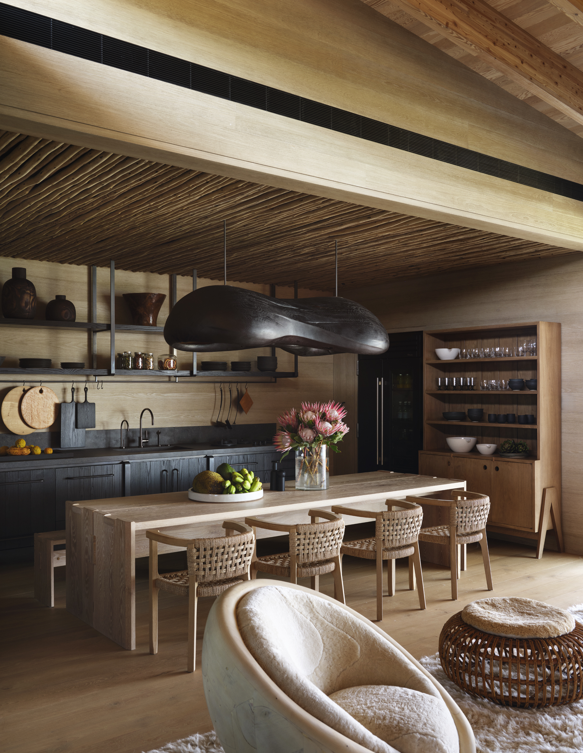A kitchen, island, dining table, and lounge share a woven-ceiling pavilion